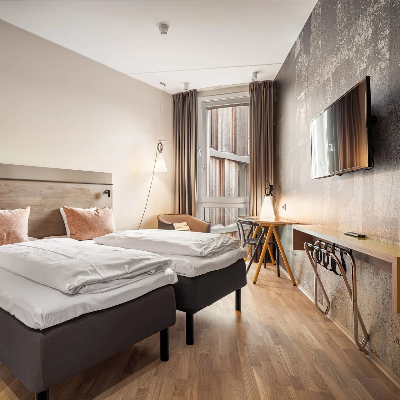 Two neatly made twin beds stand in a modern hotel room, beside a light-filled window, with wooden flooring and a wall-mounted television opposite.