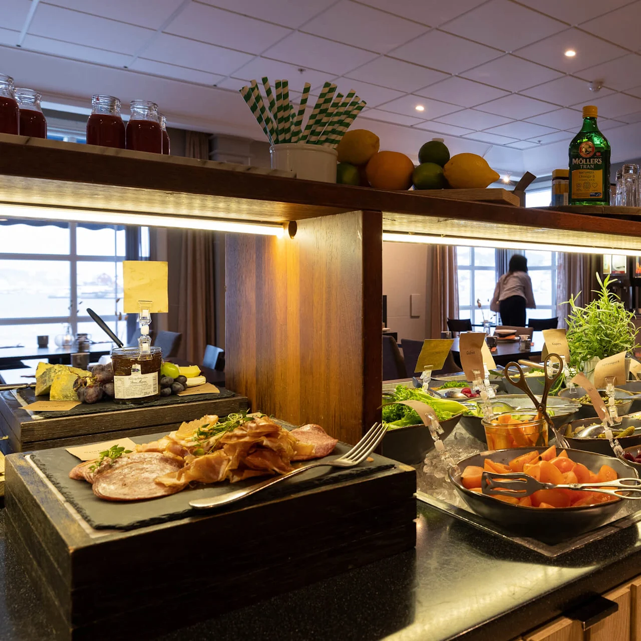 Et buffetbord viser ulike retter inkludert spekemat, frukt og salater. Ovenpå står flasker og frukt på en hylle. Et vindu og en sittende person er synlig i bakgrunnen.