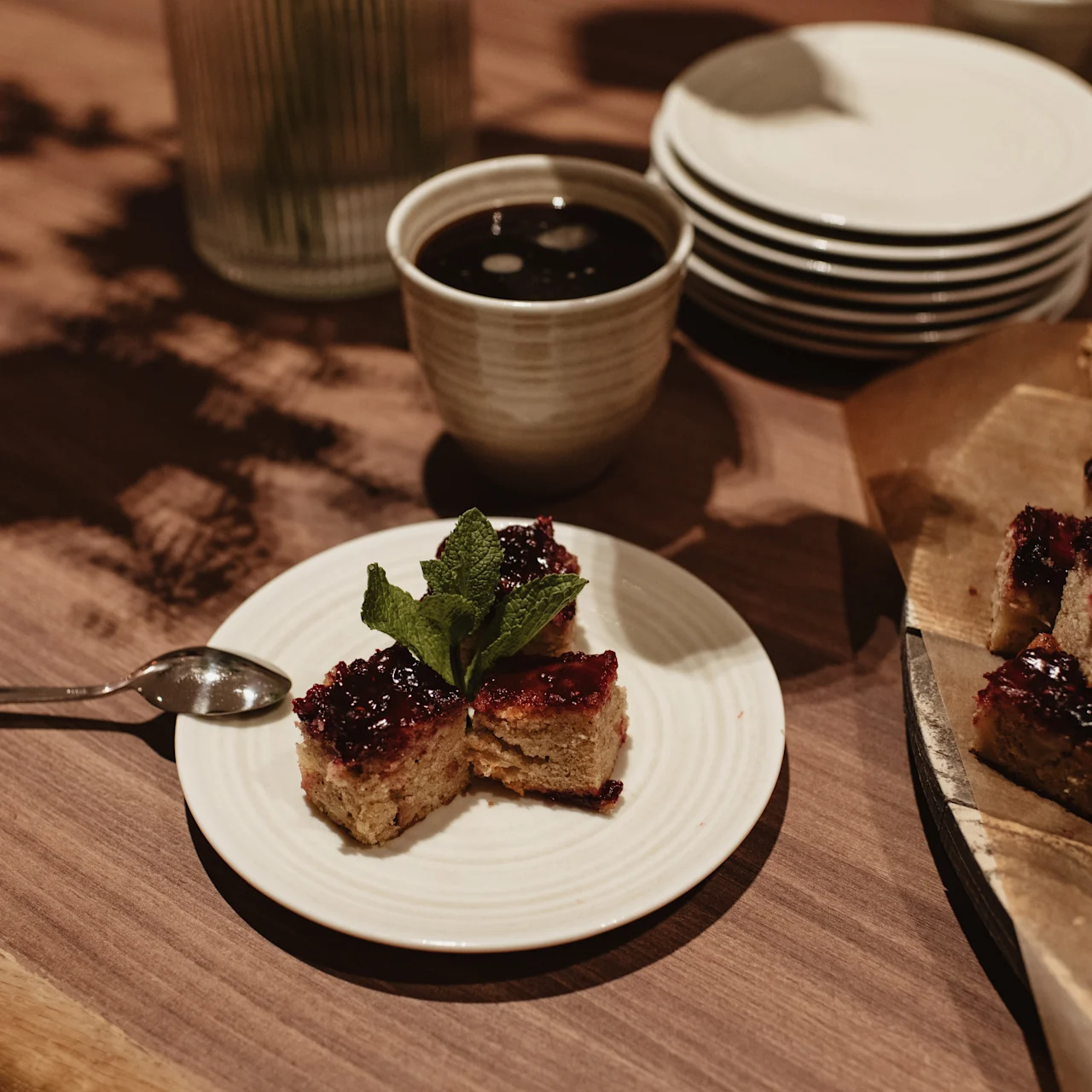 Two pieces of dessert with berry topping and mint garnish rest on a white plate beside a metal spoon on a wooden table. Nearby are stacked plates and a cup.
