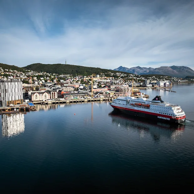 Ett stort passagerarfartyg seglar genom en hamn med en stad inbäddad mot kullar och berg under en blå himmel.