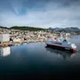 Ett stort passagerarfartyg seglar genom en hamn med en stad inbäddad mot kullar och berg under en blå himmel.