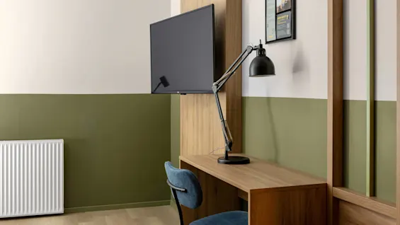 Ett skrivbord i trä med en justerbar svart lampa står mot en grön och beige vägg, bredvid en monterad TV och en inramad bild ovanför.
