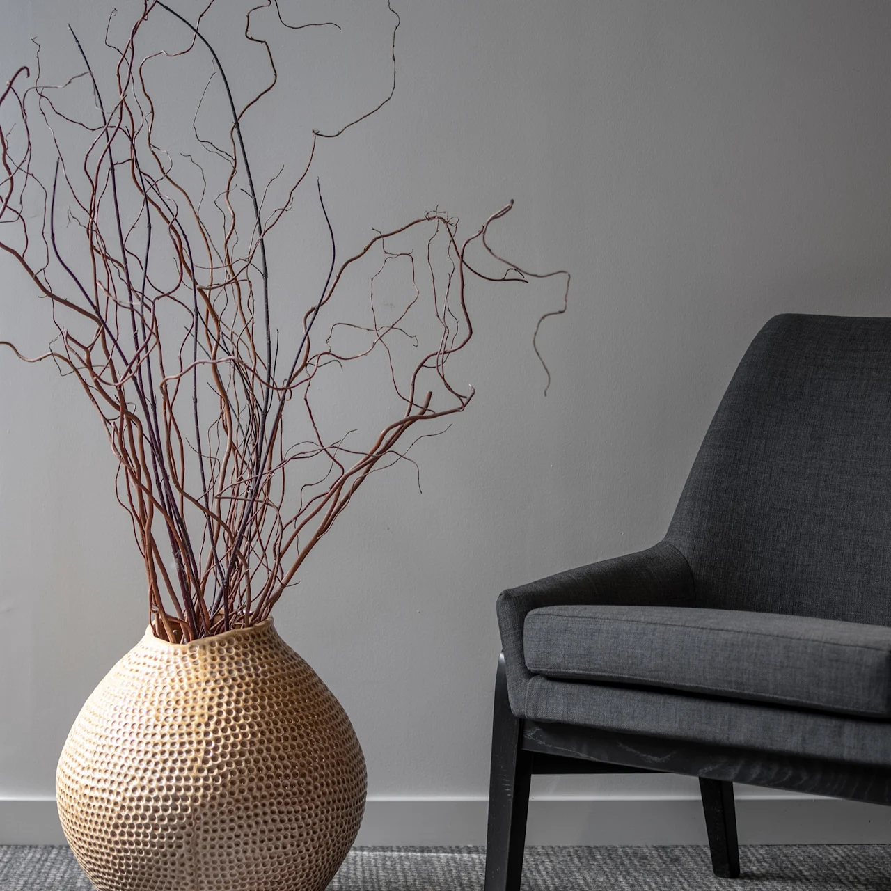 En strukturad vas håller höga, böjda grenar bredvid en grå stoppad fåtölj på en vävd matta i ett minimalistiskt, mjukt upplyst rum.