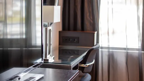 En flatskjerm-TV på et mørkt trestativ med en fjernkontroll ved siden av, står ved siden av en moderne lampe. I bakgrunnen er et skrivebord og stol nær tynne gardiner.