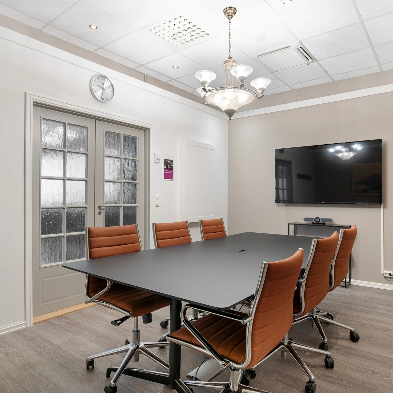A conference table with six brown chairs sits in a modern meeting room. A large TV is mounted on a beige wall. Ceiling lights illuminate the space. Clock shows 9:30. On the wall, text: "hegna."
