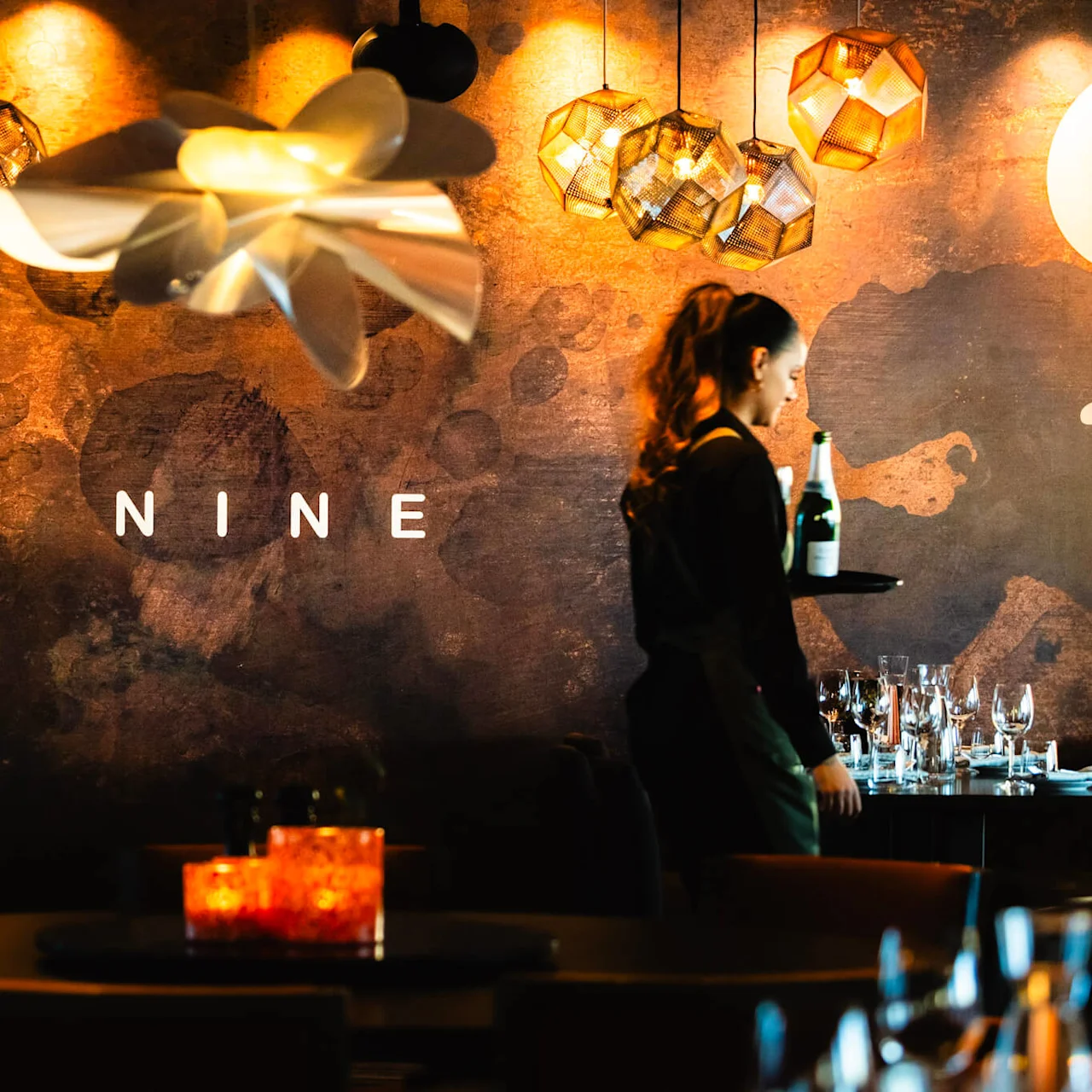 En servitör bär en bricka med en flaska i en dämpat upplyst restaurang. Dekorativa lampor hänger ovanför. På väggen står texten "NINE".