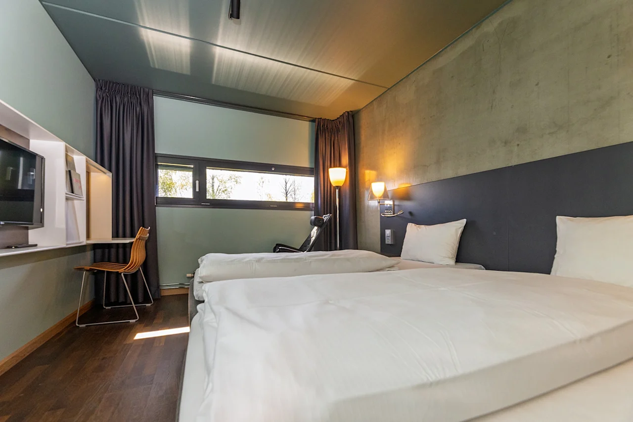Två prydligt bäddade sängar i ett modernt hotellrum med trägolv. Ett skrivbord med stol står vid ett fönster med gardiner, upplyst av vägglampor.