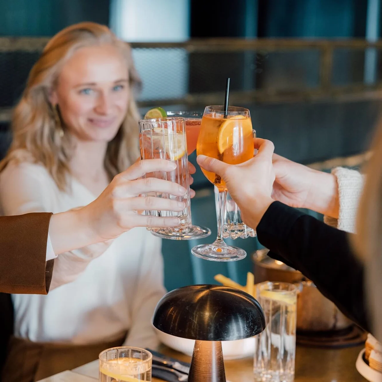 Glas med drikkevarer, der klinkes sammen, holdt af mennesker, der sidder ved et spisebord og fejrer i en varmt oplyst restaurant.