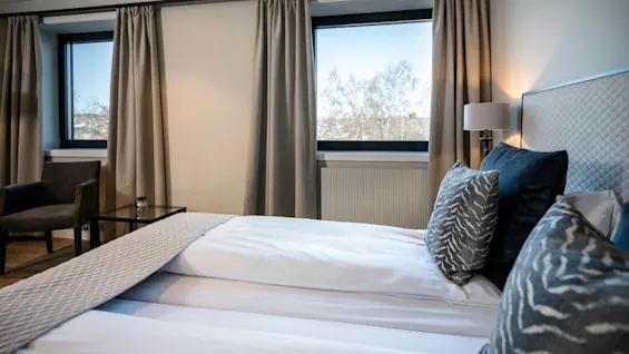 En pænt redt seng med mønstrede puder står i et moderne, lyst rum med store vinduer, beige gardiner og en lampe ved siden af; en stol og et bord er i nærheden.