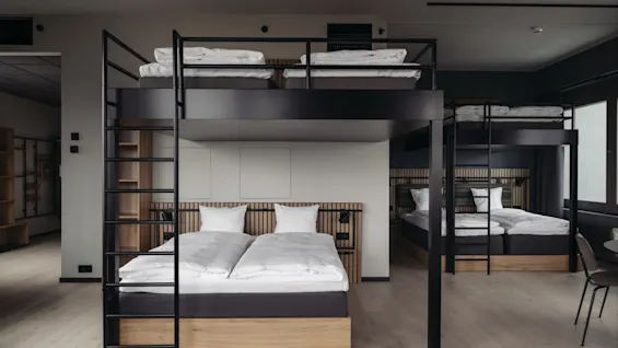 Two sets of bunk beds with wooden frames, neatly made with white linens, stand in a modern, minimalist room with light wood floors and minimal decor.
