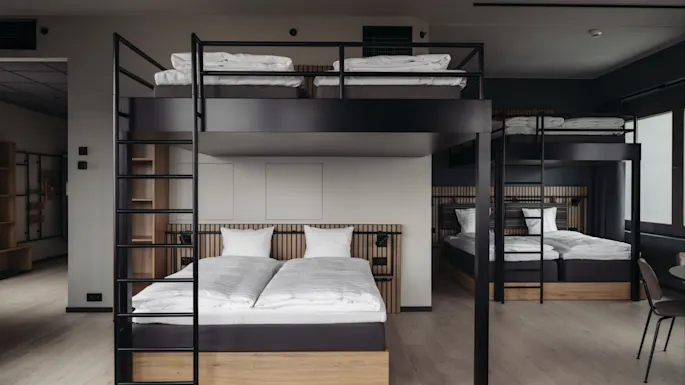 Two sets of bunk beds with wooden frames, neatly made with white linens, stand in a modern, minimalist room with light wood floors and minimal decor.