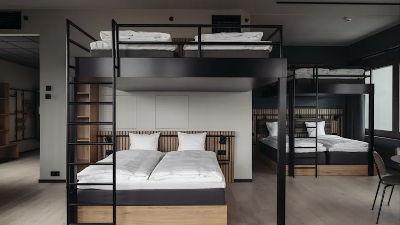 Two sets of bunk beds with wooden frames, neatly made with white linens, stand in a modern, minimalist room with light wood floors and minimal decor.