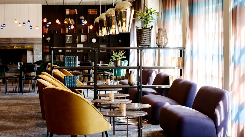 Gule stole vender mod runde borde i en hyggelig lounge, med bogreoler og planter. Lilla sofaer er placeret ved solbeskinnede vinduer med tynde gardiner.