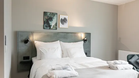 Viihtyisä hotellihuone, jossa siististi pedattu sänky, valkoiset liinavaatteet ja kaksi taideteosta sängynpäädyn yläpuolella. Puhtaat pyyhkeet ovat valmiina.