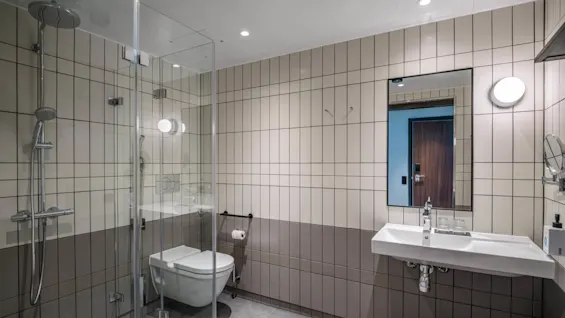 Et moderne bad har en glassinnrammet dusj, veggmontert toalett og bredt servant under et speil. Beige fliser dekker veggene, og skaper et rent og moderne rom.