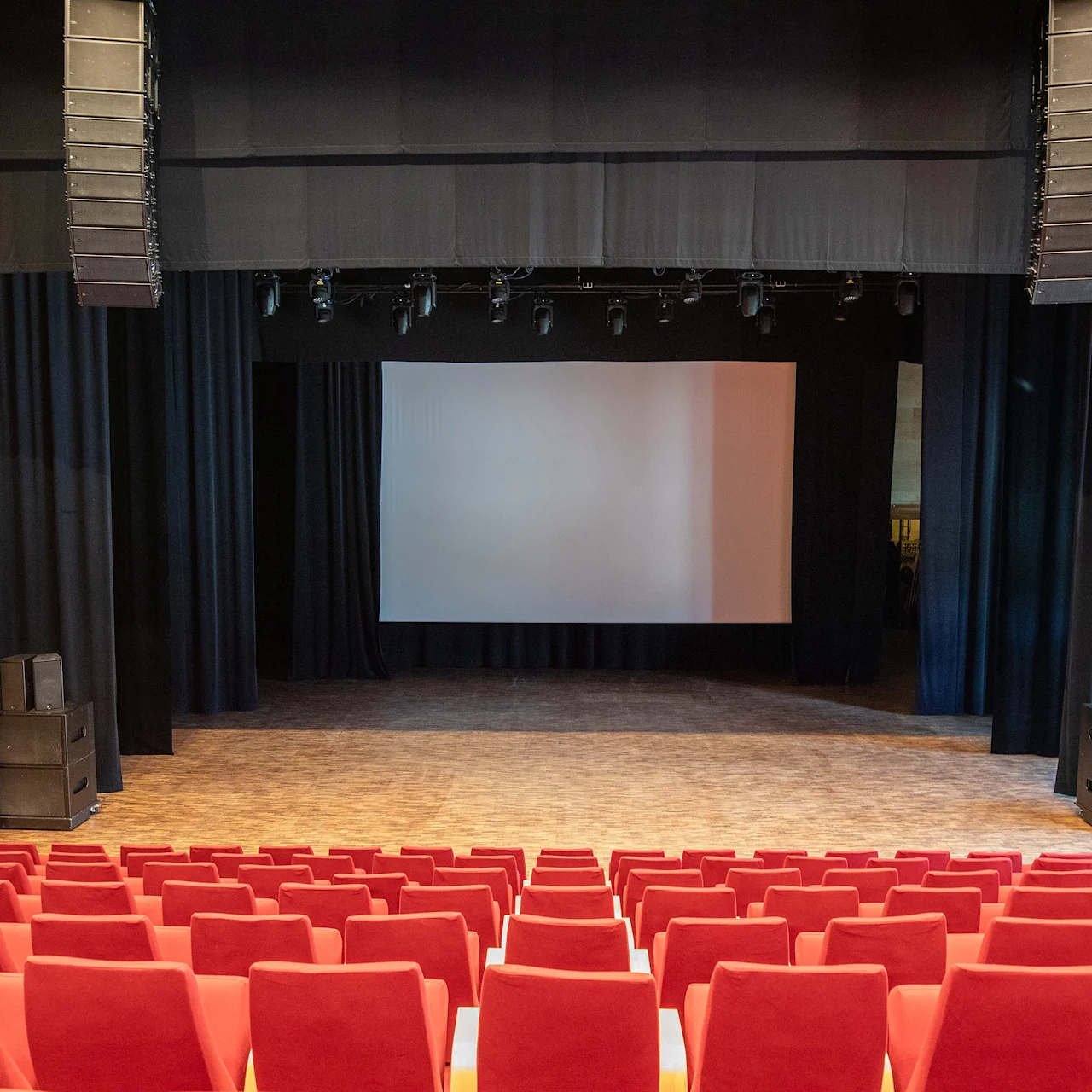 En scene med et tomt hvitt lerret er omgitt av svarte gardiner, sett fra rader med røde teaterstoler i et stort auditorium.