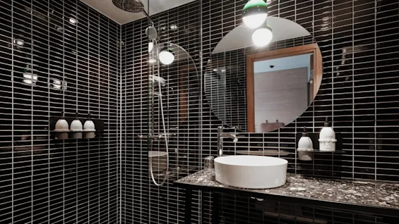 A circular mirror above a sink is mounted on a black-tiled bathroom wall with bottles labeled "Shampoo," "Shower," and "Lotion" on the wall shelf nearby.