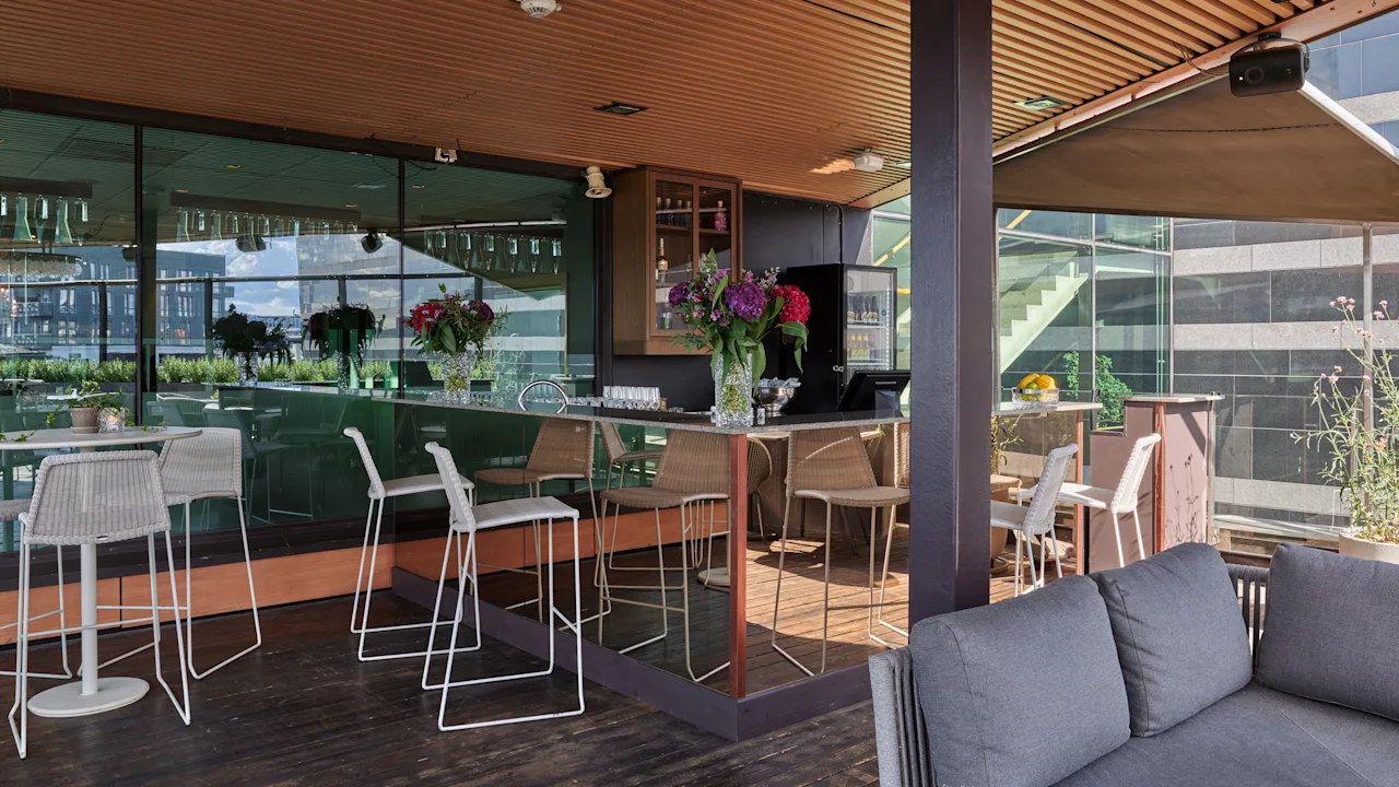Bar med vita stolar och bord placerade på ett trädäck. Glasväggar och tak, dekorativa växter och färgglada blommor kompletterar den utomhus urbana miljön med närliggande byggnader synliga.