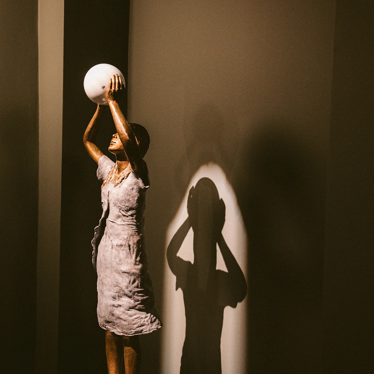A sculpture of a woman in a dress raises a spherical object, casting a shadow on a dark wall in a dimly lit space.