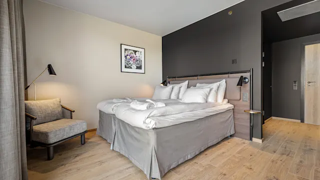 En nyoppredd dobbeltseng står i et moderne hotellrom ved siden av en polstret stol. Myk belysning og nøytrale farger. Veggkunst over sengen tilfører dekor.