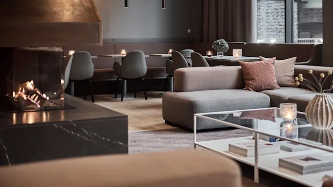 Moderne salong med peis, grå sofaer og et stilfullt salongbord. Stearinlys og minimalistisk dekor skaper en koselig atmosfære med store vinduer i bakgrunnen.