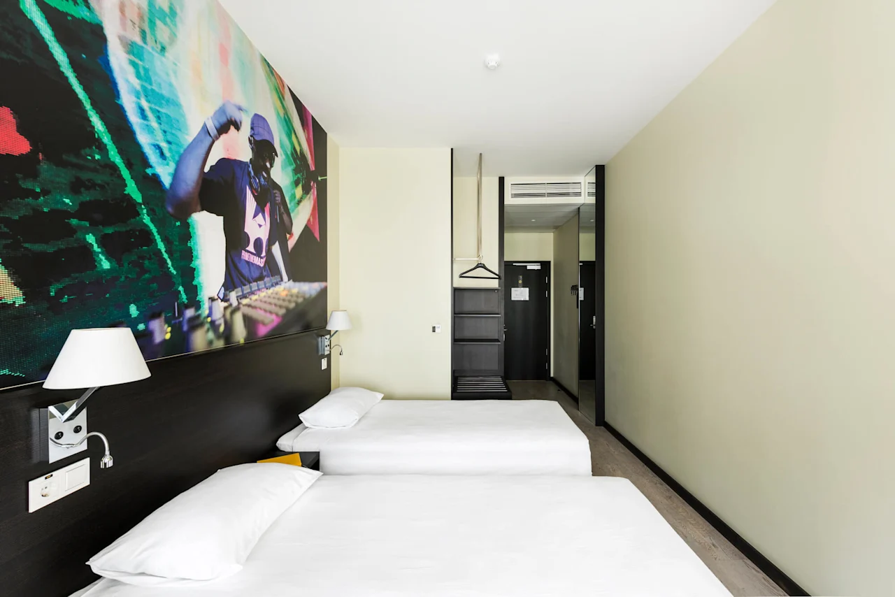 Two beds with white linens are located in a modern hotel room. A colorful DJ mural is on the wall, and a hanger and shelf are near the entrance.