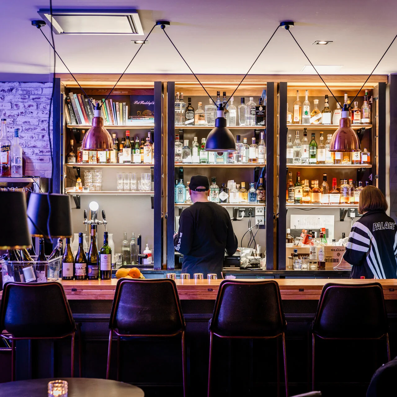 Bardisk fylt med flasker; en bartender og en kunde interagerer. Den koselige atmosfæren har stoler, hengende lamper og dekorative murvegger.