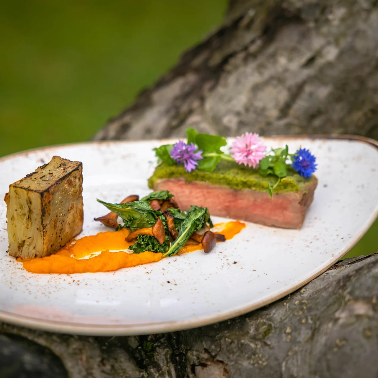 En gourmetret med stekt kjøtt pyntet med spiselige blomster og urter, sammen med stekte grønnsaker og oransje saus på en rustikk tallerken, mot en naturlig utendørs bakgrunn.