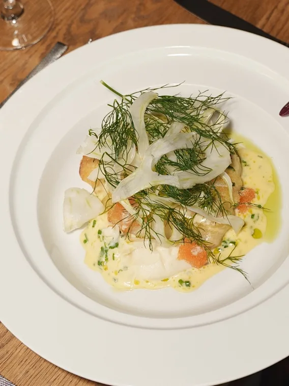 En hand serverar en elegant fiskrätt med fänkål och örter på en vit tallrik vid ett dukat träbord.