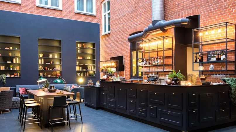 An elegant indoor bar area with stools, shelves with books and bottles, set against exposed brick walls and dark decor.