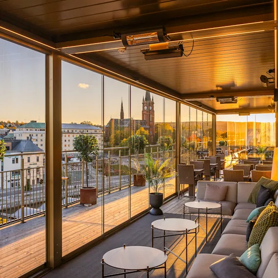 Inglasad takterrass i solnedgång med bekväma soffor, små bord och panoramautsikt över staden.