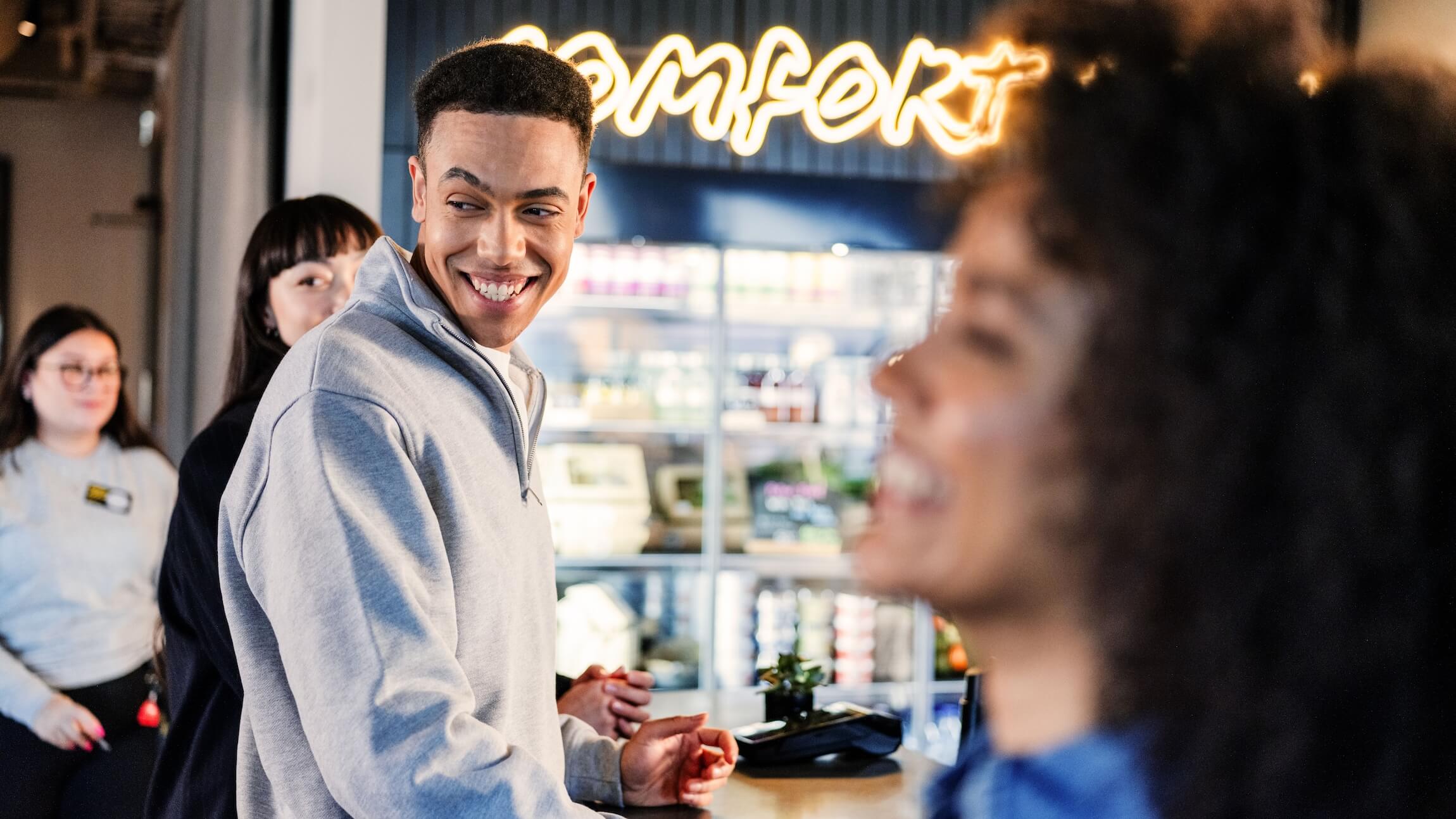 En person i en ljusgrå tröja ler mot två personer i ett modernt café med en neonskylt "Comfort" i bakgrunden.