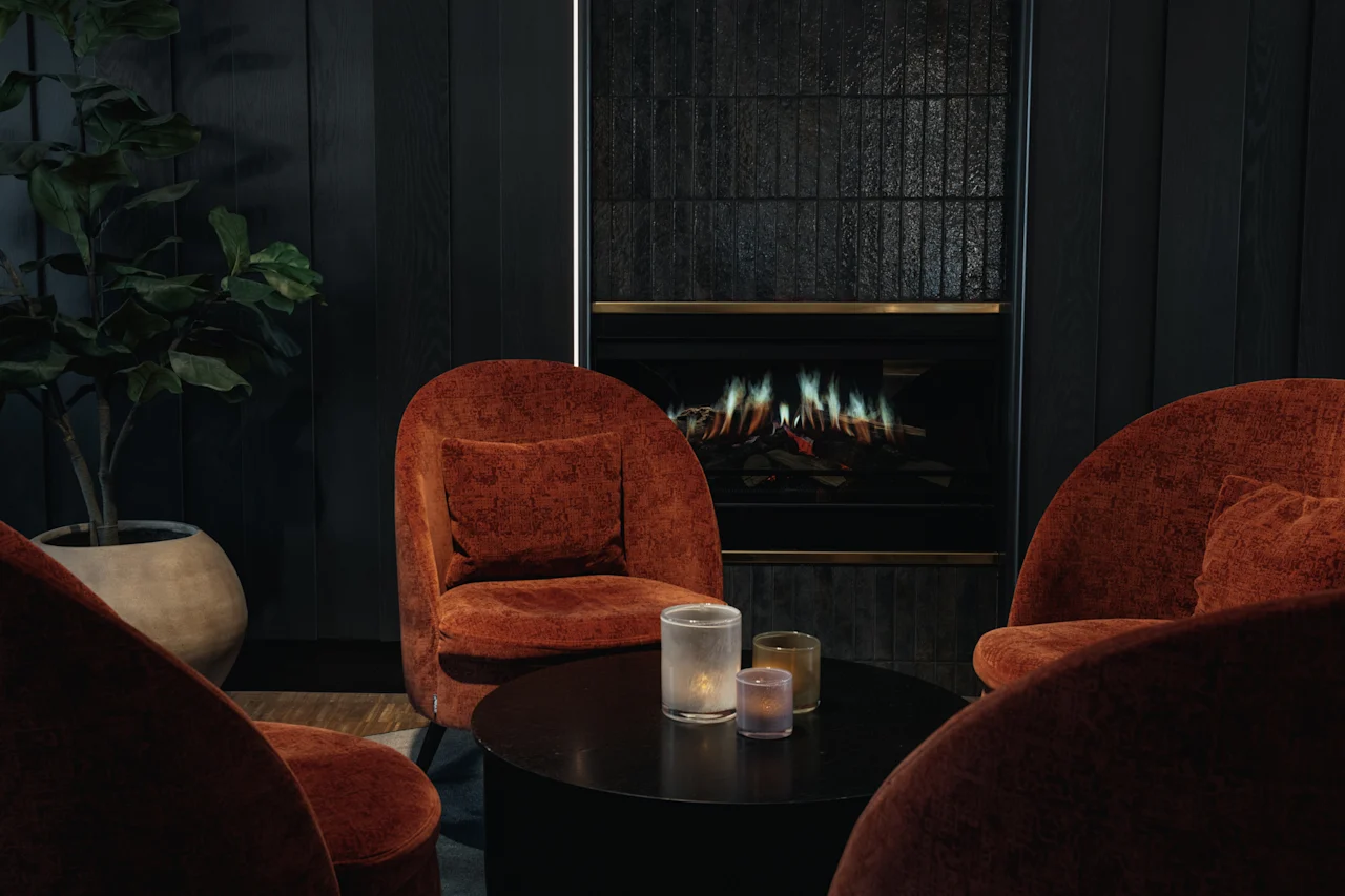 Et hyggeligt loungeområde med rustfarvede lænestole omkring et sofabord med stearinlys, mod en mørk væg med pejs.