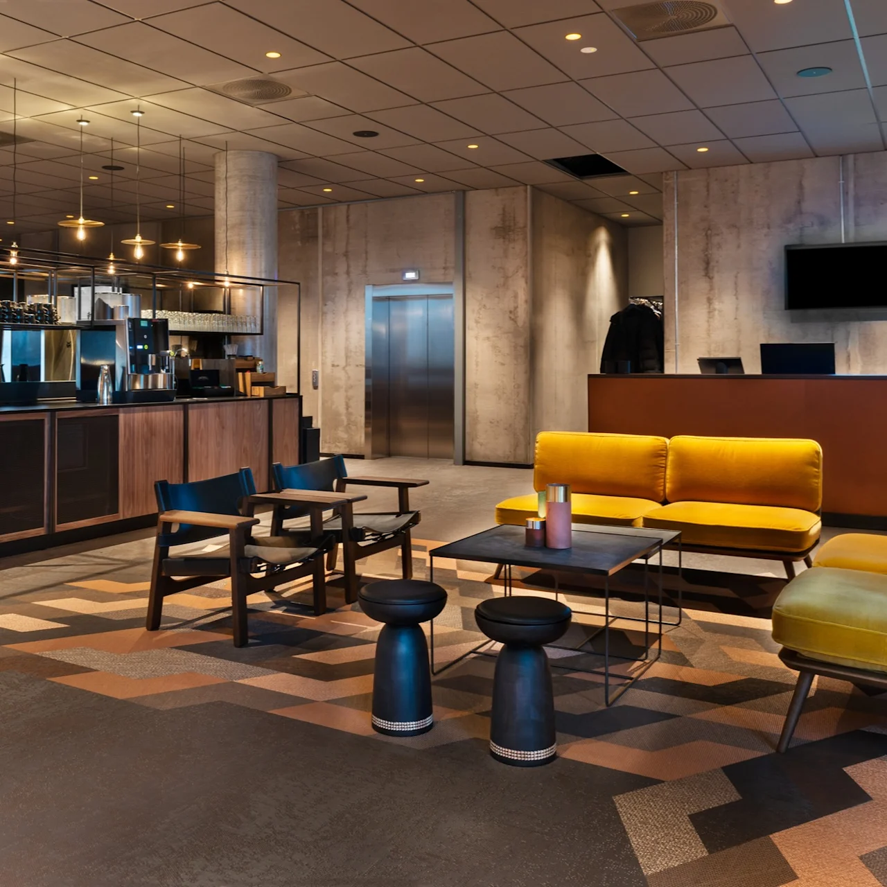 En moderne lounge har gule sofaer og svarte stoler rundt et lavt bord. Til venstre er en bar med kaffestasjon. I bakgrunnen ses en heis og en resepsjon.