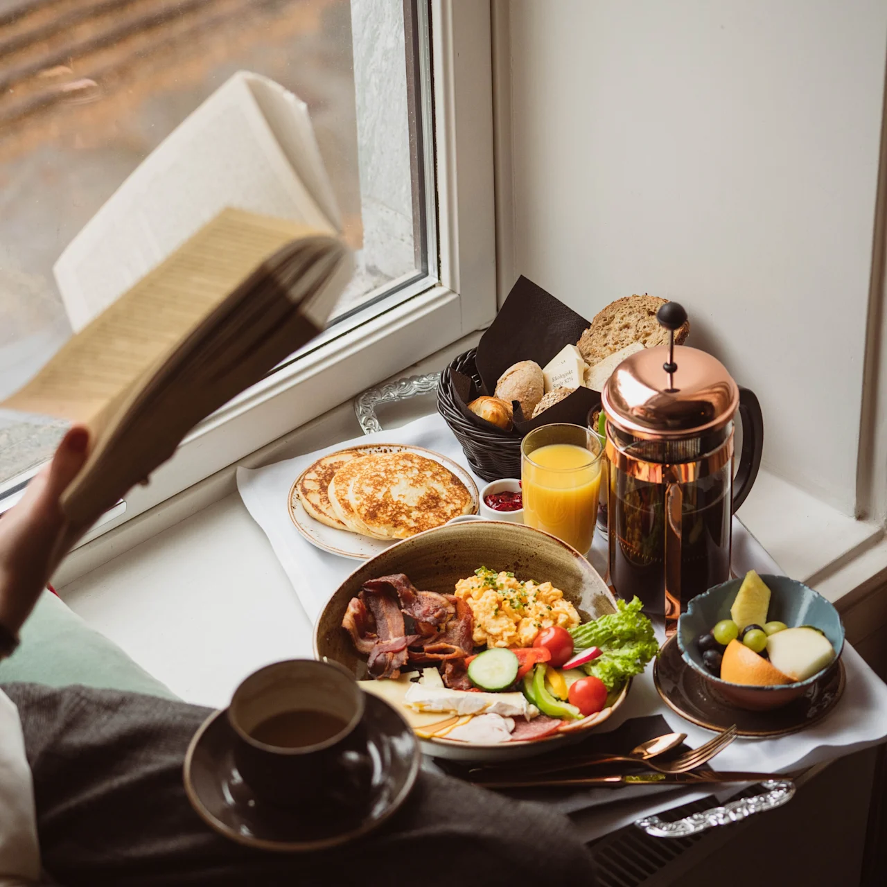 A breakfast tray with scrambled eggs, bacon, pancakes, assorted breads, orange juice, and a French press. Person reading a book beside a window with a city view.