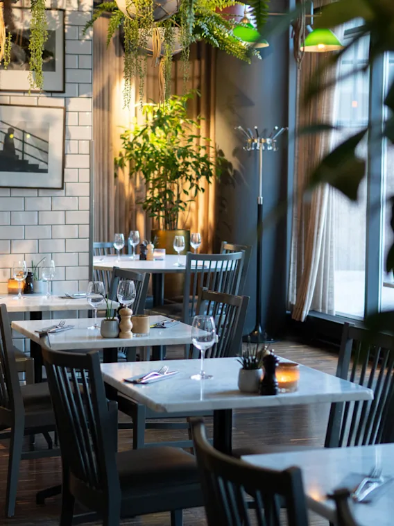 Bord med hvite topper, dekket med glass og bestikk, står i en koselig restaurant pyntet med planter og innrammede bilder, opplyst av varm belysning.