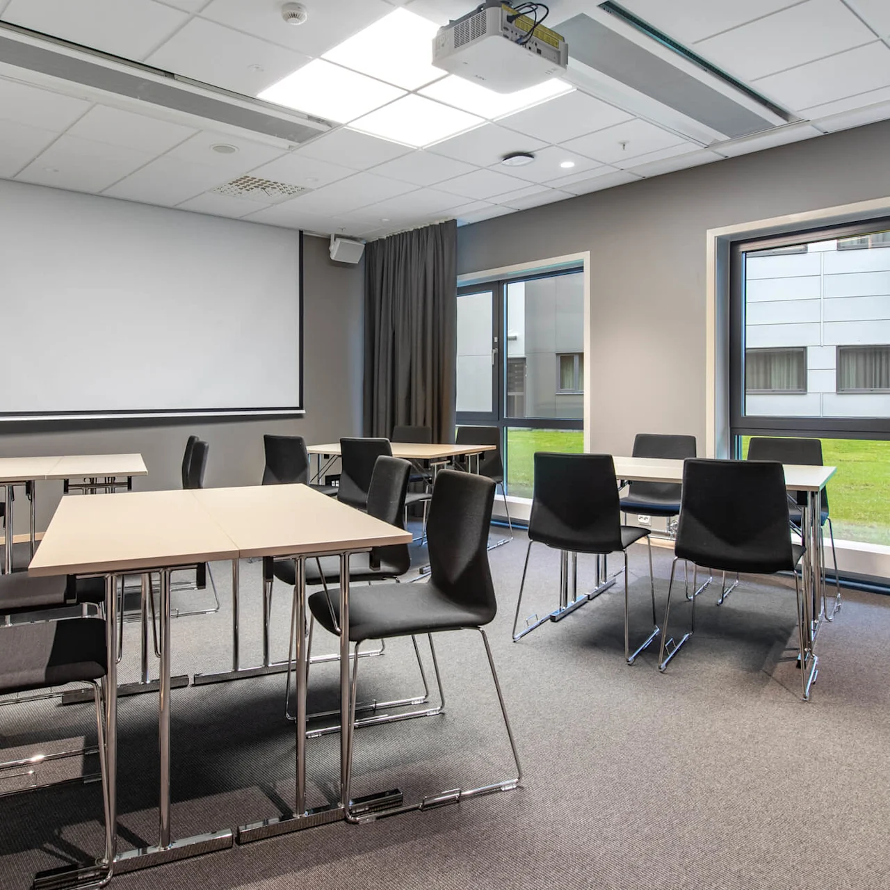 A classroom with black chairs and light wooden tables is arranged under a ceiling projector and screen. Large windows provide natural light, showing a grassy outdoor area.