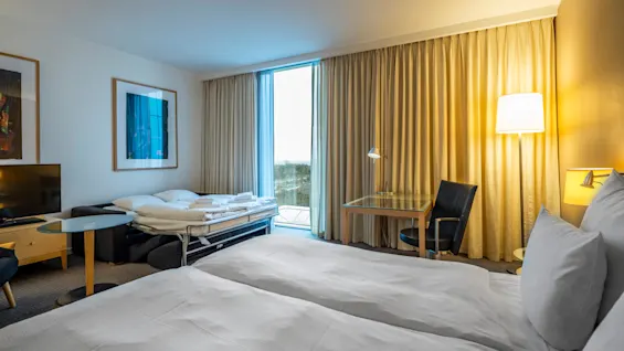 Ett hotellrum innehåller en snyggt bäddad säng och en ihopvikt bäddsoffa. Ett skrivbord och stol står under en tänd lampa vid fönstret, med inramad konst på väggen.