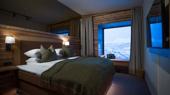 A cozy bedroom features a neatly made bed with green cushions and blanket. Large window reveals a snowy mountain landscape outside. Wooden paneling and soft lighting create a warm atmosphere.
