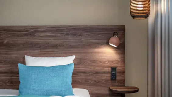 A bed with a turquoise pillow rests against a wooden headboard. A reading lamp is attached to the headboard. A woven ceiling light and curtains frame the cozy room.