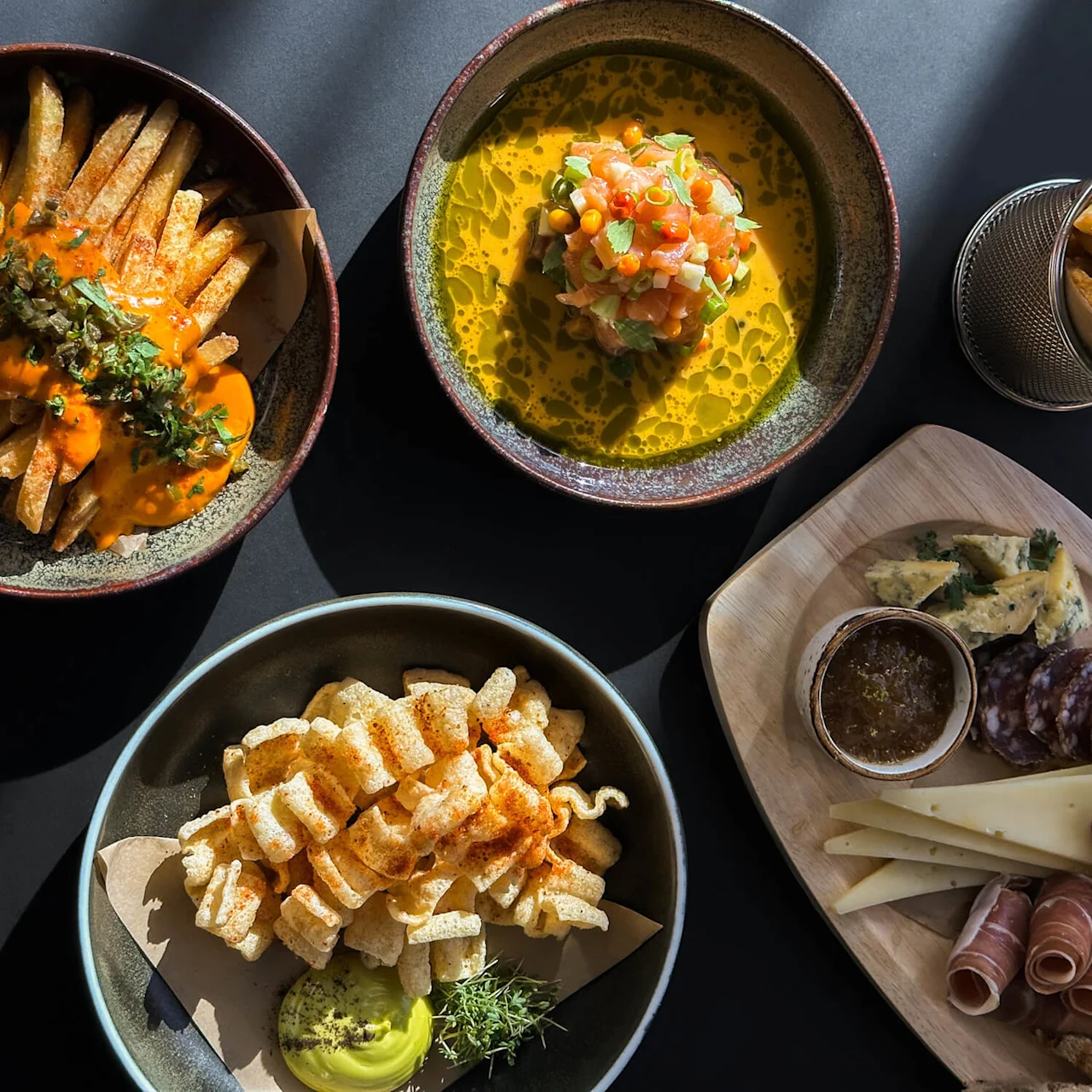 A variety of gourmet dishes on a table: fries with sauce, a dish with diced toppings in broth, fries in a basket, pork rinds with dip, and a charcuterie board with cheese, meat, grapes, and spreads.
