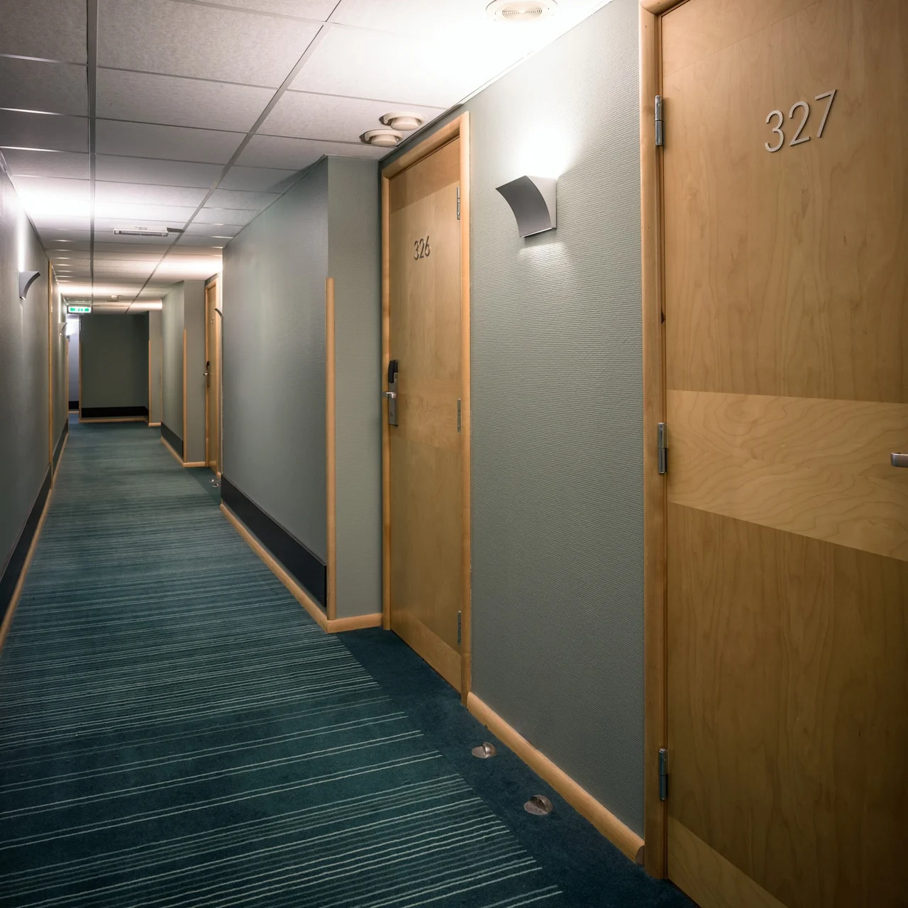 En korridor med dörrar numrerade "326" och "327" leder till rum. Mjuk belysning lyser upp gröna väggar och en mönstrad matta, vilket skapar en lugn hotellkorridor.