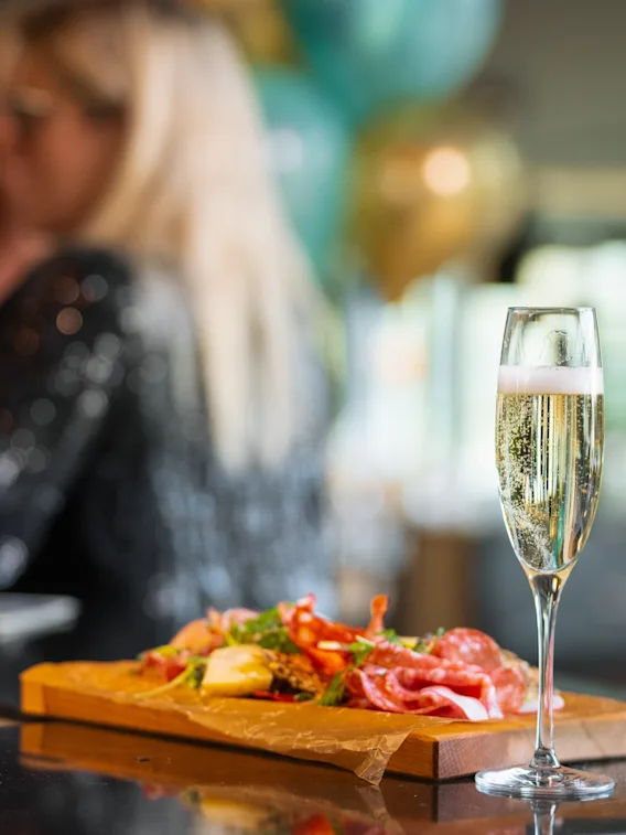 Champagneglass på disk, ledsaget av et charcuteriebrett toppet med kjøtt, grønnsaker og urter. Uklart bakgrunn avslører en person som holder et lignende glass i en festlig setting.