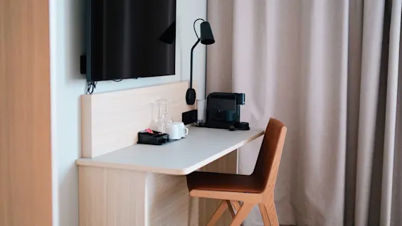 Desk holds coffee machine and cups; modern chair sits beside; TV mounted above; lamp attached. Beige curtains frame one side, creating a cozy hotel room setting.