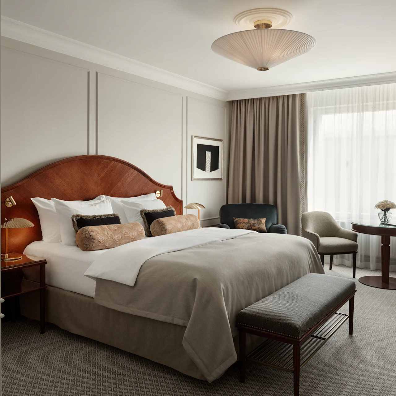 A neatly-made double bed with pillows and a wooden headboard sits in a hotel room. Nearby, there's a table with two chairs, a window with curtains, and a stylish ceiling light.