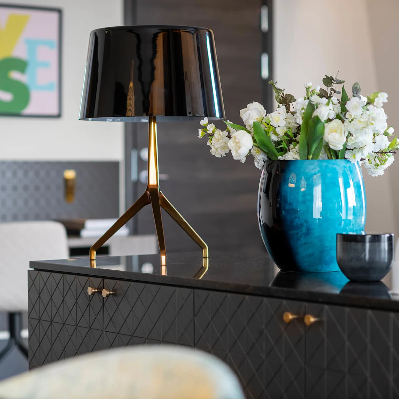 A black lamp stands on a dark patterned cabinet with gold handles. Beside it, a blue vase holds white flowers. In the background, abstract art reads "YES."