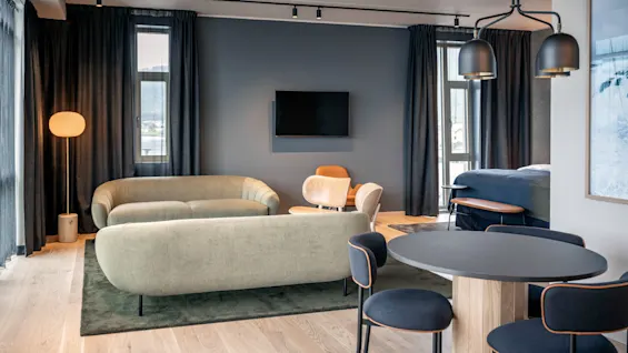 En moderne stue har et rundt spisebord med sorte stole, to grønne sofaer og et vægmonteret TV. Blød belysning og store vinduer skaber en hyggelig atmosfære.