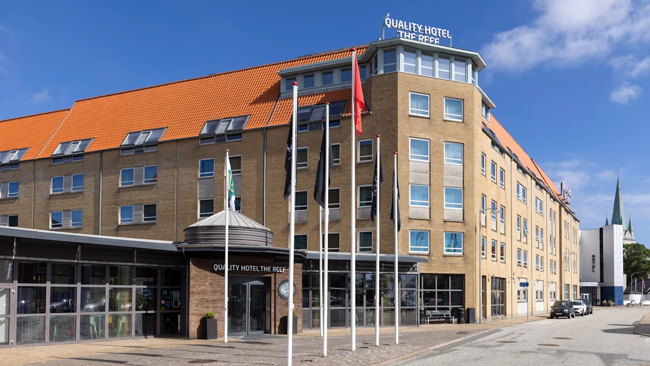 Et fleretagers hotel med rødt tegltag står på en solrig gade, med skilte der viser "QUALITY HOTEL THE REEF" på facaden. Flere flag er placeret foran.
