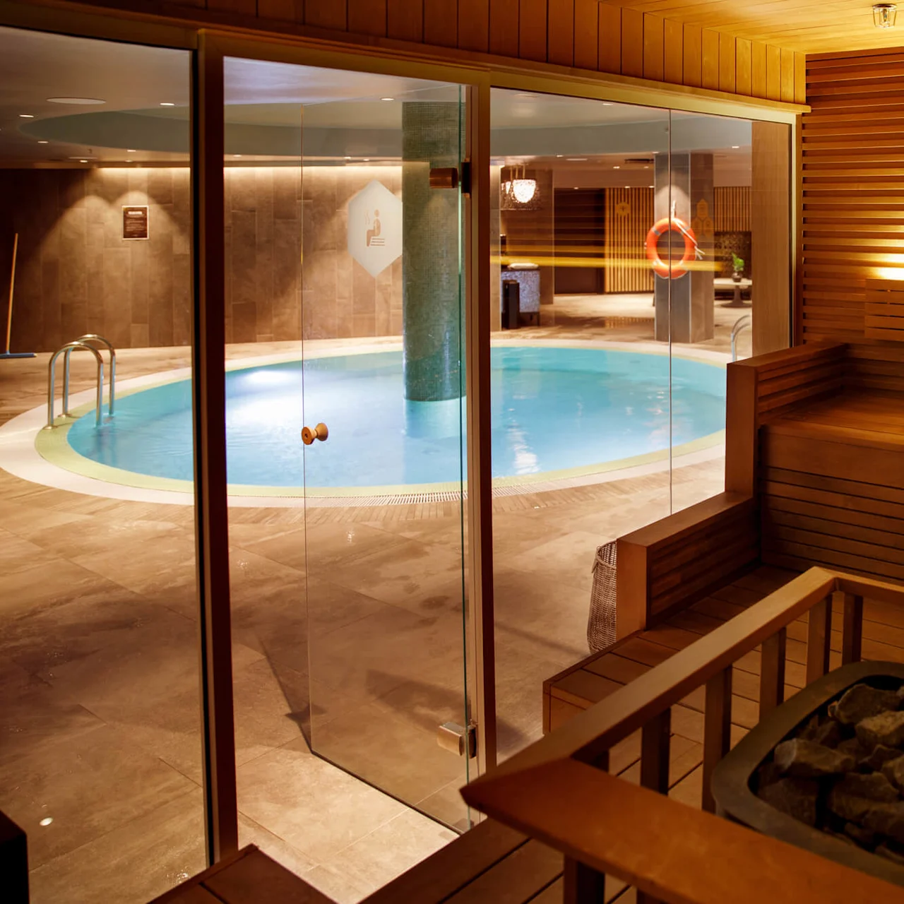 Sauna interior with wooden benches and rocks, viewed through glass doors looking into a tiled indoor pool area. A pool ladder is visible with dim lighting surrounding.