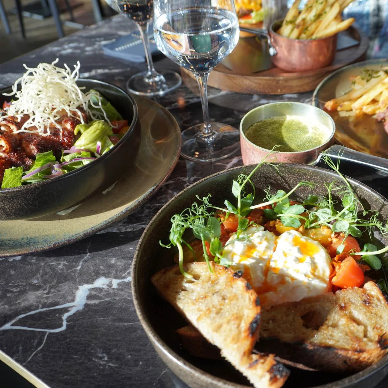 A table with three plates of food, including a salad with crispy garnish, a steak with fries, and a dish with toast and vegetables. Glasses of water and wine accompany.
